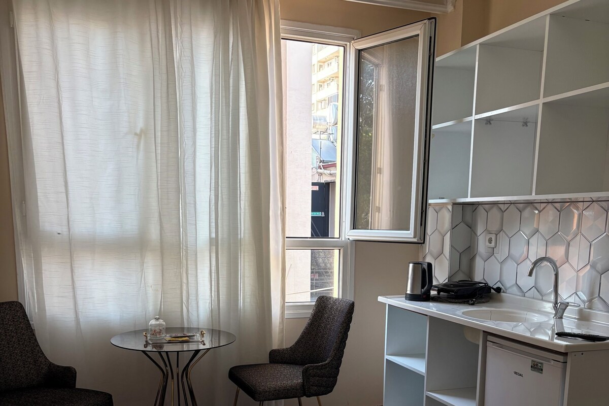 A hotel room with a bed, a small table, two chairs, a kitchenette, and a window with curtains.