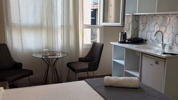 Ein Hotelzimmer mit einem Bett, einem kleinen Tisch, zwei Stühlen, einer Küchenzeile und einem Fenster mit Vorhängen.