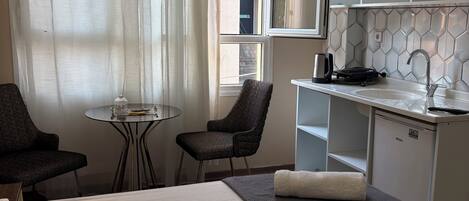 Hotellihuone, jossa on sänky, pieni pöytä, kaksi tuolia, pieni keittiönurkkaus ja ikkuna, jossa on verhot.