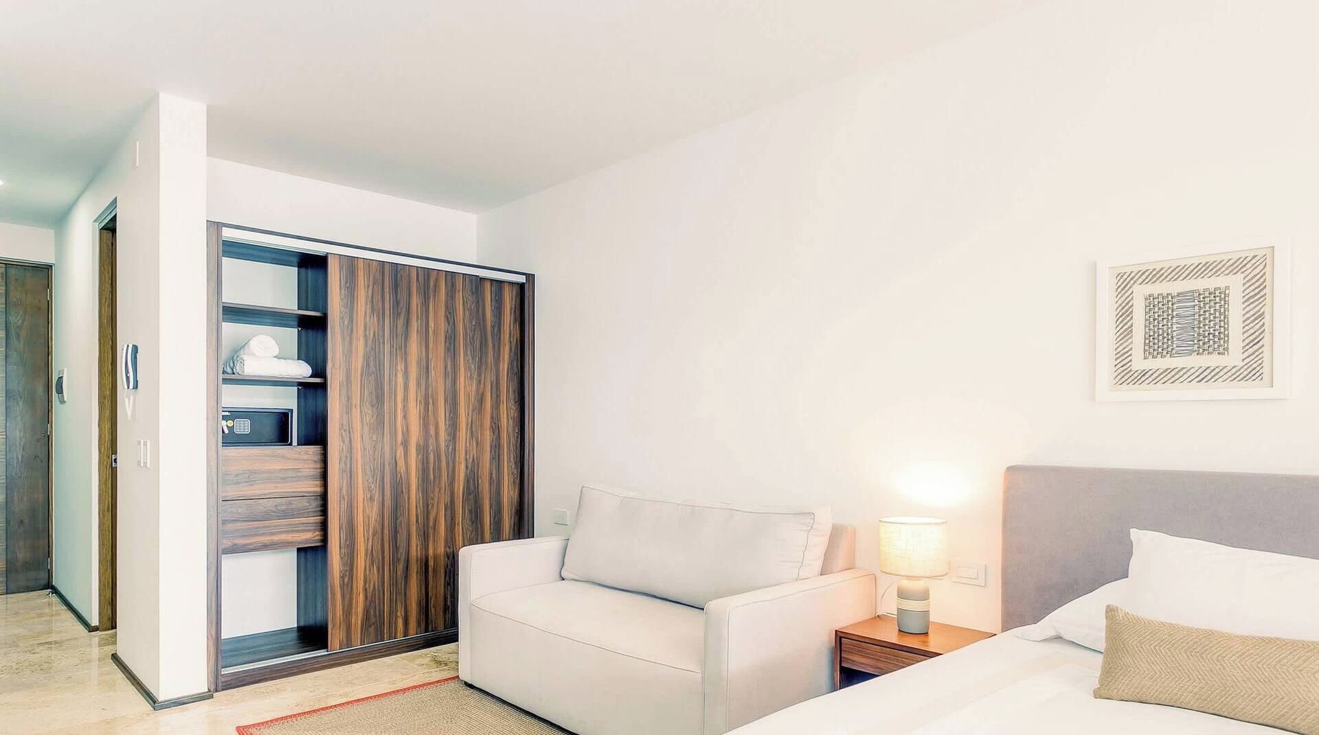 Une chambre d’hôtel avec un lit, une table de chevet avec une lampe, un canapé blanc, une armoire et une porte donnant sur une autre pièce.