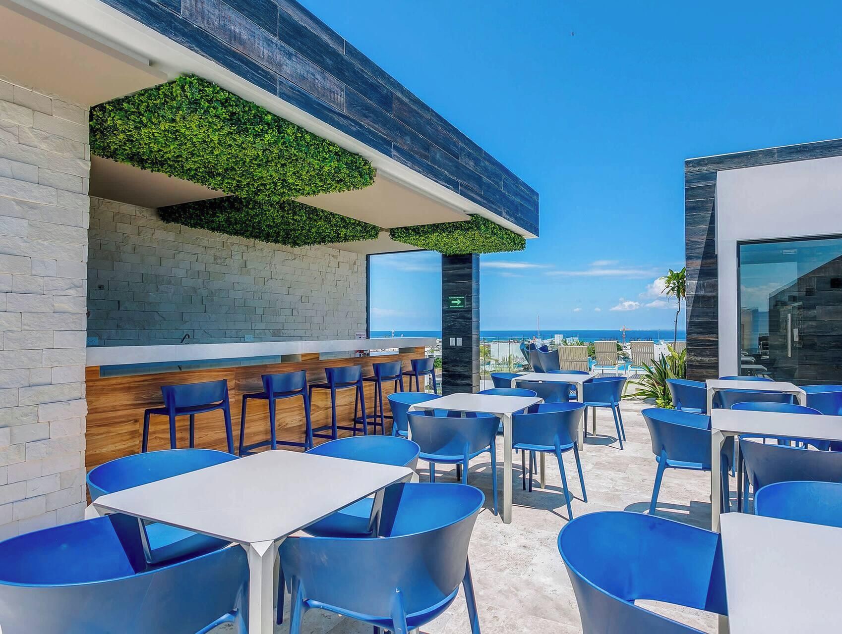 An outdoor seating area with blue chairs and tables, a view of the ocean, and a modern building facade with a brick wall and greenery.