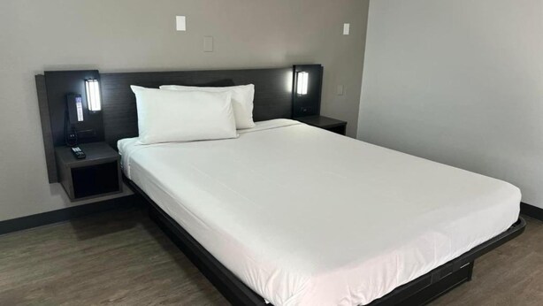 Cama bem arrumada com roupa de cama branca, cabeceira escura e abajures laterais.
