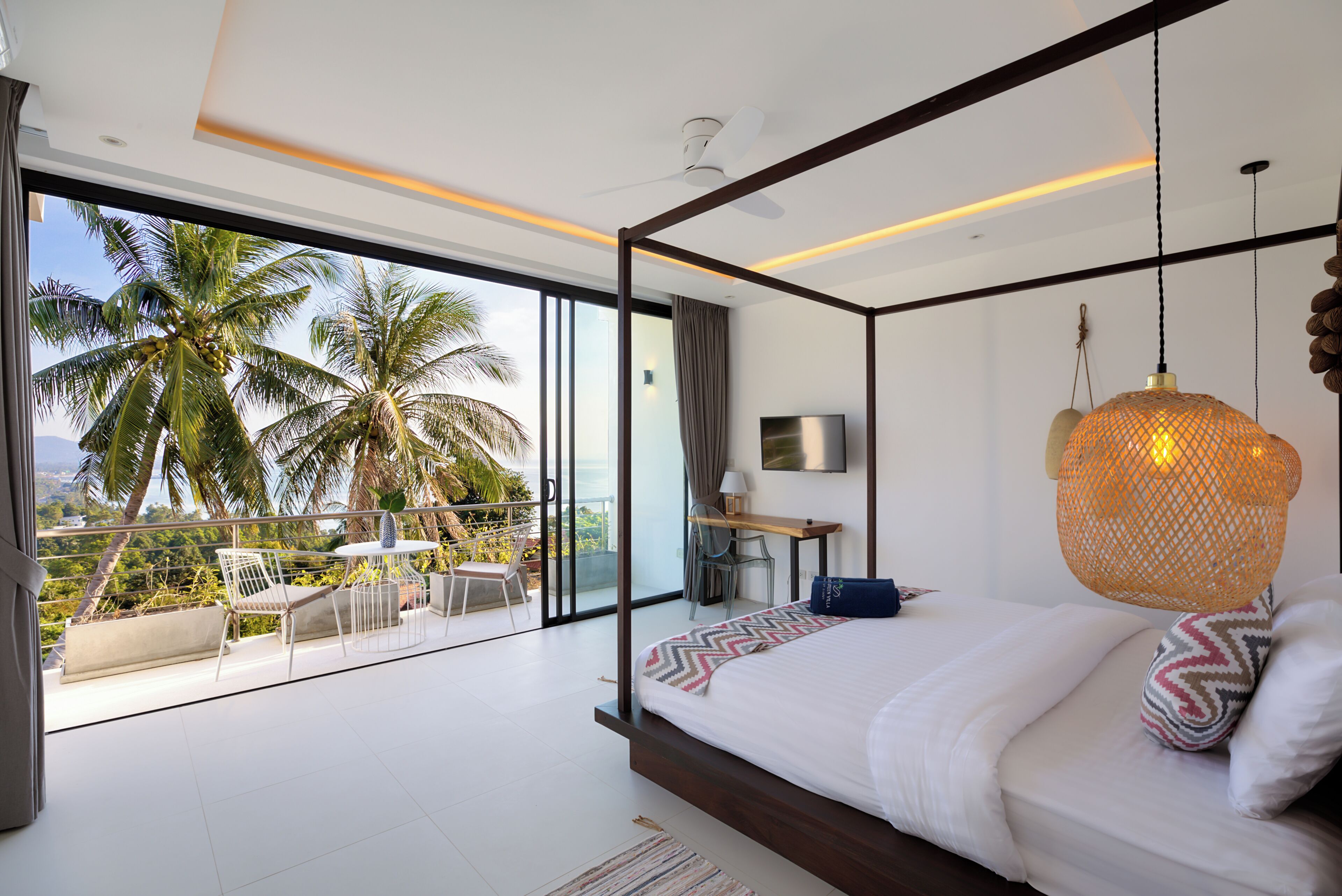 A modern bedroom with a large bed, a desk, and a balcony with palm trees.