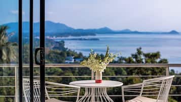 A balcony with a table and two chairs, overlooking a coastal landscape with mountains and a body of water.
