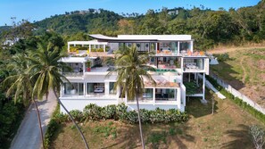 A multi-level modern house with a pool, surrounded by palm trees and a hilly landscape. - 4 Bedroom Seaview Tropical Eco-friendly Villa (Koh Samui)