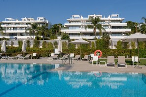 A swimming pool with lounge chairs and umbrellas, surrounded by a fence and palm trees. - Cateleya Residence by Time & Home (Estepona)