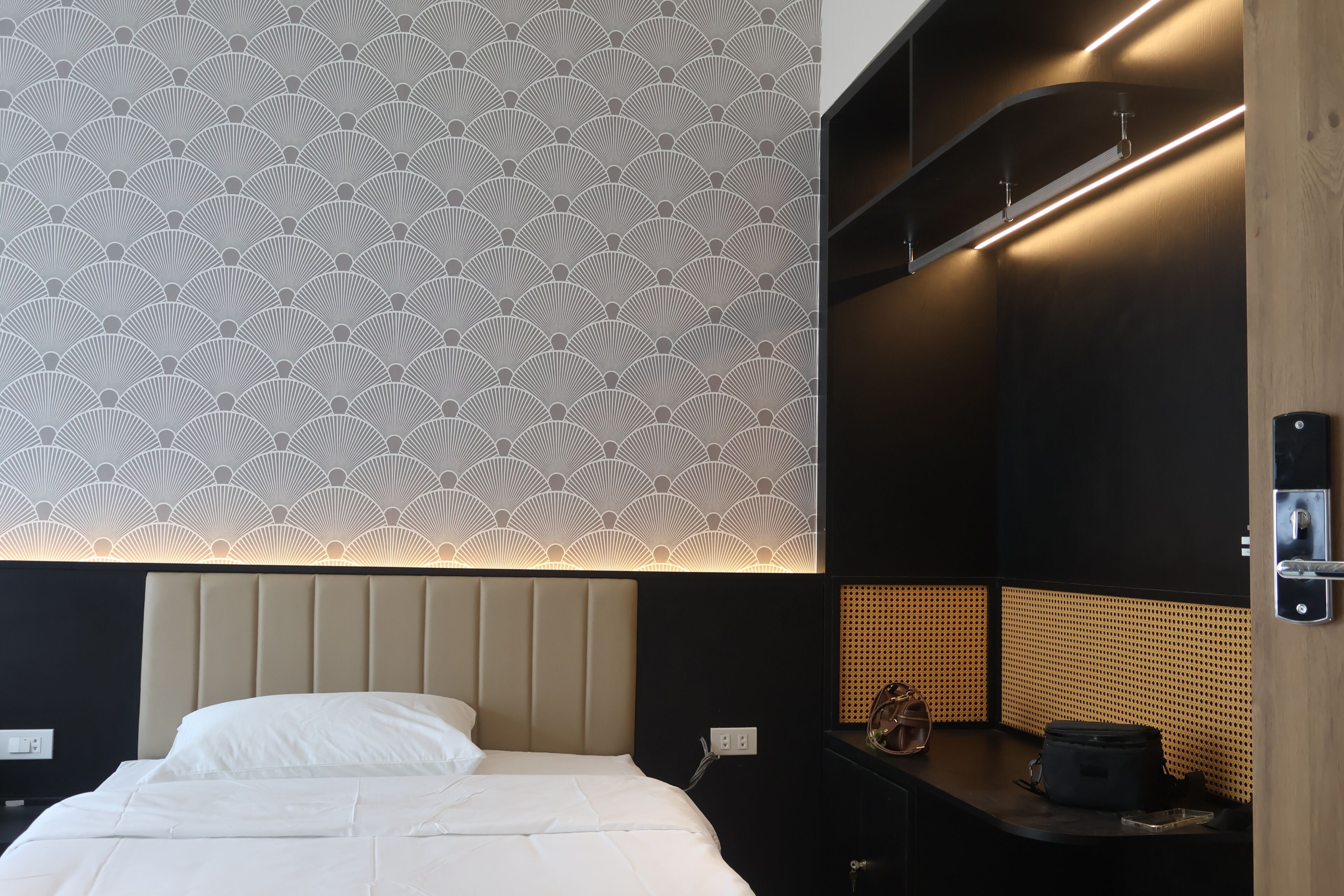 A hotel room with a patterned wall, a bed with white bedding, a desk with a lamp, and a wall-mounted shelf.
