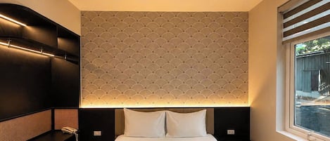 A modern hotel room with a large bed, a bedside table, a built-in bench, a patterned wall, and a window with blinds.