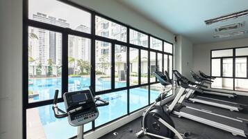 A gym with treadmills, a stationary bike, and a rowing machine, with a large window overlooking a swimming pool and high-rise buildings.