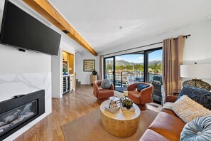 Ein modernes Wohnzimmer mit Kamin, großem Fernseher und Blick auf Berge und Boote durch die Glasfenster.