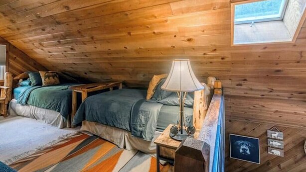 Un dormitorio acogedor en el ático con paredes de madera, un tragaluz, dos camas, una mesita de noche y una lámpara.