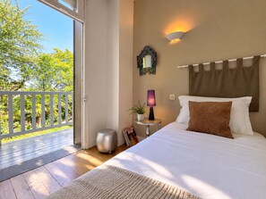 Ein Schlafzimmer mit einem Bett, einem Nachttisch, einer Lampe, einem Wandspiegel und Blick ins Grüne.