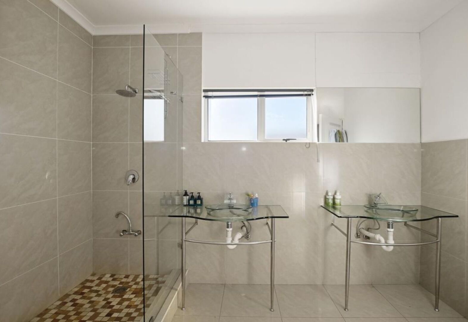 Een moderne badkamer met een douchecabine van glas, twee glazen wastafels en een raam boven de wastafels.