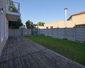 En træterrasse der fører ud til en græsplæne med et metalhegn, en skorsten og en lille legeplads.