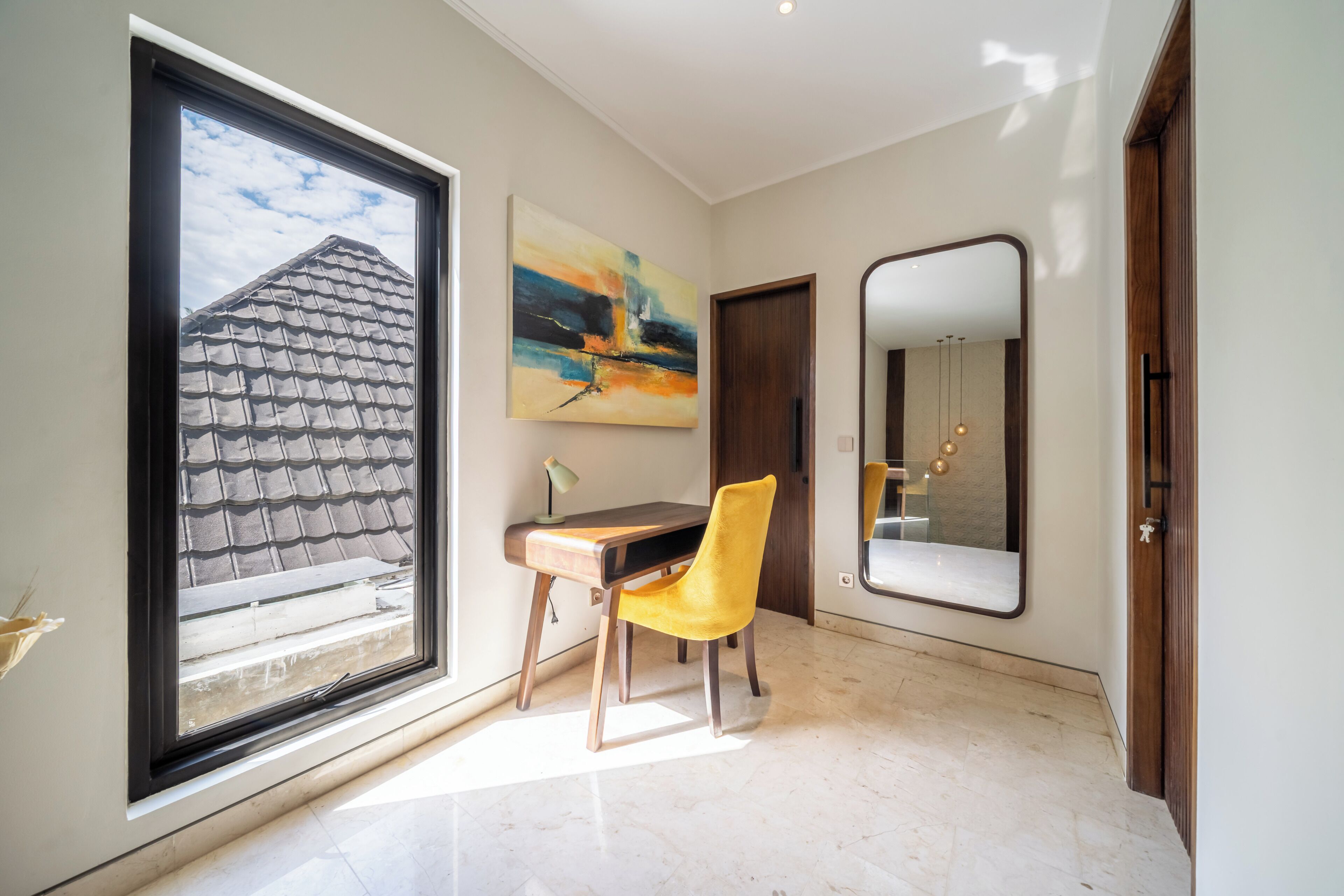 A room with a large window, a desk, a chair, a painting, and a mirror.