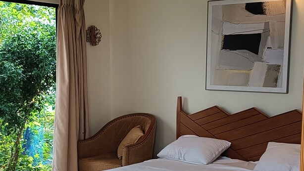 A bedroom with a bed, a ceiling fan, a wall-mounted light, a chair, and a window with curtains. - you can enjoy the mountains, rivers greenery & modern luxury resort together. (Rangamati)