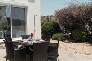 An outdoor dining area with wicker chairs and a table set for two, a grill, and a view of a hillside.