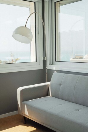 Ein Zimmer mit einem hellgrauen Sofa, einer Stehlampe und einem Fenster mit Meerblick.
