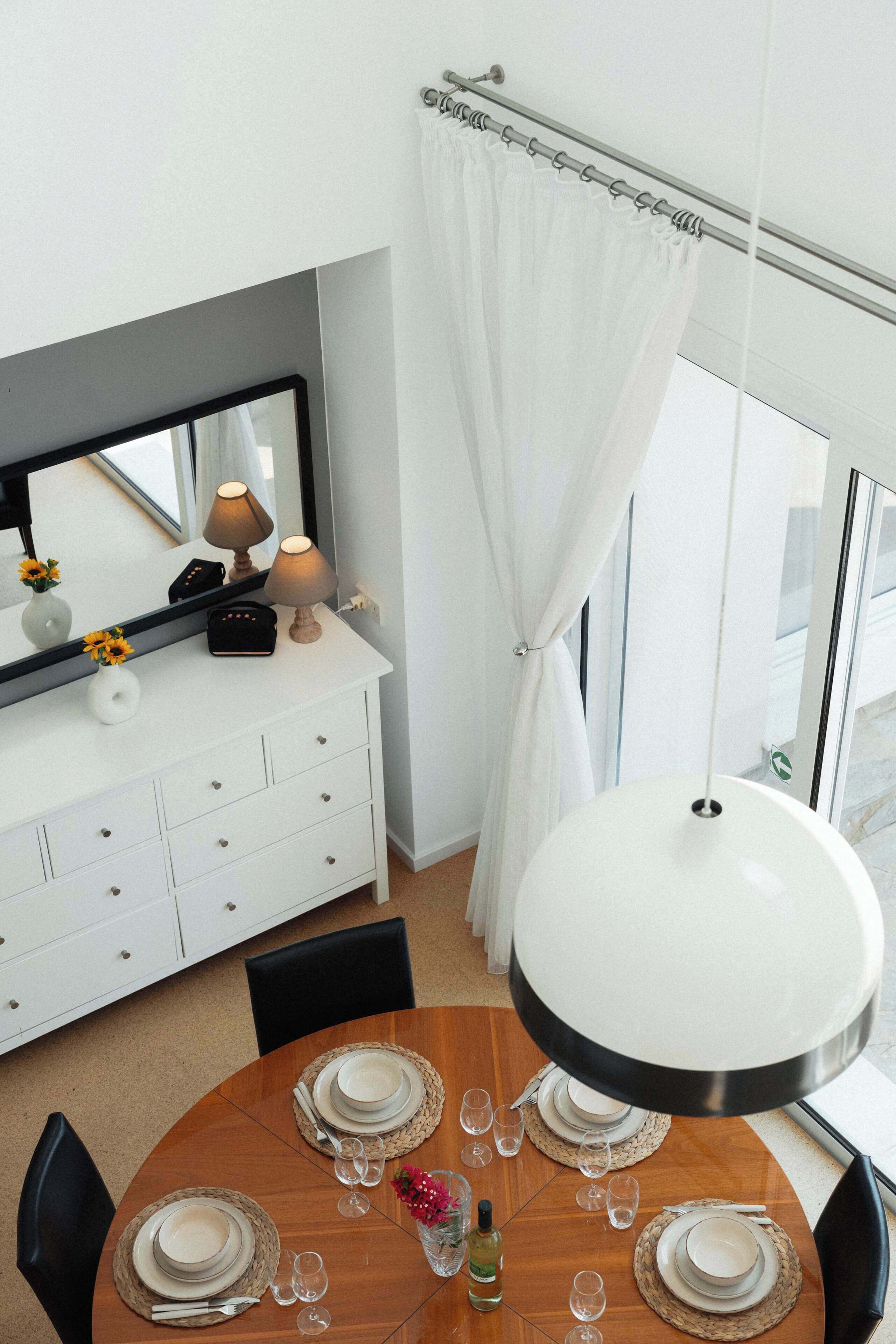 Ein Essbereich mit einem runden Tisch, gedeckt für vier Personen, einer weißen Konsole mit Spiegel, Lampen und einer Vase mit Sonnenblumen.
