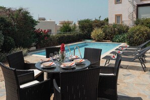 A poolside dining area with a table set for four, surrounded by wicker chairs, and a pool with lounge chairs and a palm tree. - 3BR Panoramic Sea View Villa with Private Pool (Pissouri)
