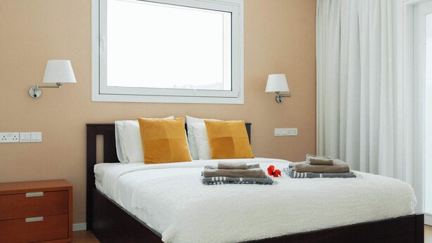 A neatly made bed with white bedding and two yellow pillows, a wooden nightstand with a drawer, and two wall-mounted lamps. - 3BR Panoramic Sea View Villa with Private Pool (Pissouri)
