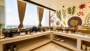 A buffet setup with various food stations, a city view through large windows, and a wall decorated with plant illustrations.
