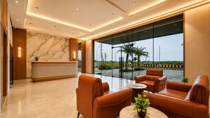A modern lobby with a reception desk, leather seating, a marble floor, and large windows offering a view of palm trees and a road. - Aarambh by The Sky Imperial Jamnagar (Jamnagar)