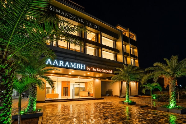 Un hotel llamado 'AARAMBH by The Sky Imperial' con una fachada moderna, ventanas amplias y un área de entrada pavimentada con palmeras.