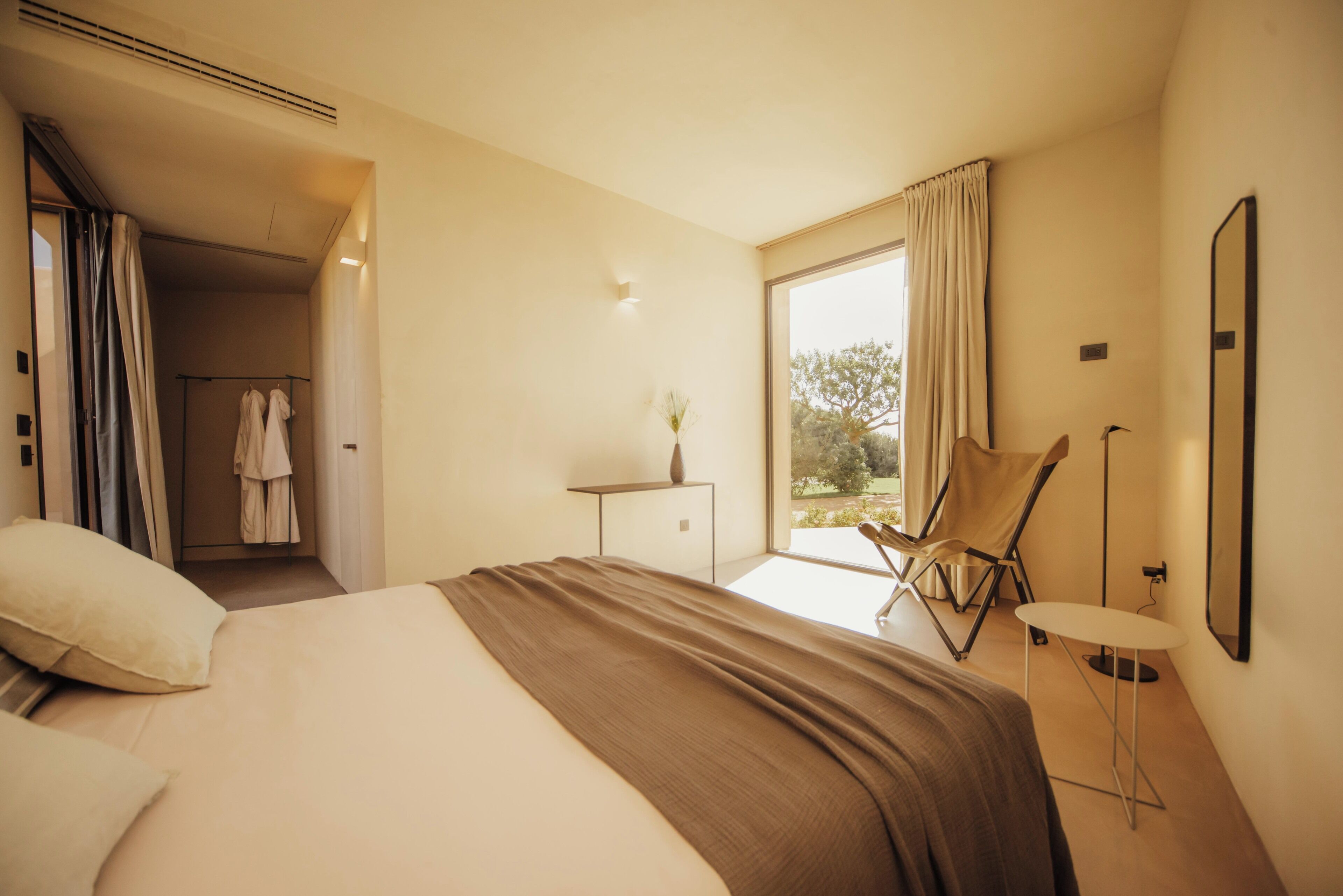 Une chambre d’hôtel moderne dotée d’un grand lit, d’un coin salon avec un fauteuil et une petite table, et offrant une vue sur l’extérieur.