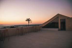 A rooftop terrace with a view of the city at sunset, featuring a palm tree and several chairs.