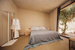 Un dormitorio moderno con una cama grande, una mesita de noche con lámpara, una bata colgada en un perchero y una ventana con vistas a árboles.