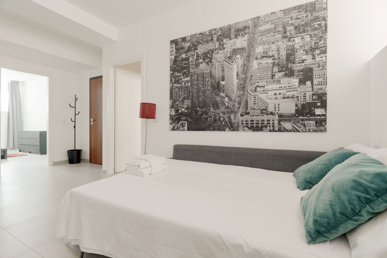 A modern hotel room with a large bed, a cityscape artwork on the wall, a red lamp, and a view of the hallway.