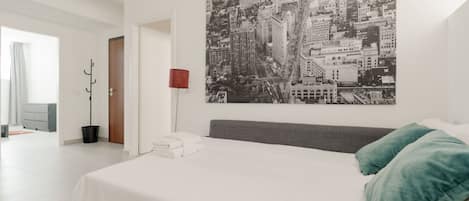 A modern hotel room with a large bed, a cityscape artwork on the wall, a red lamp, and a view of the hallway.