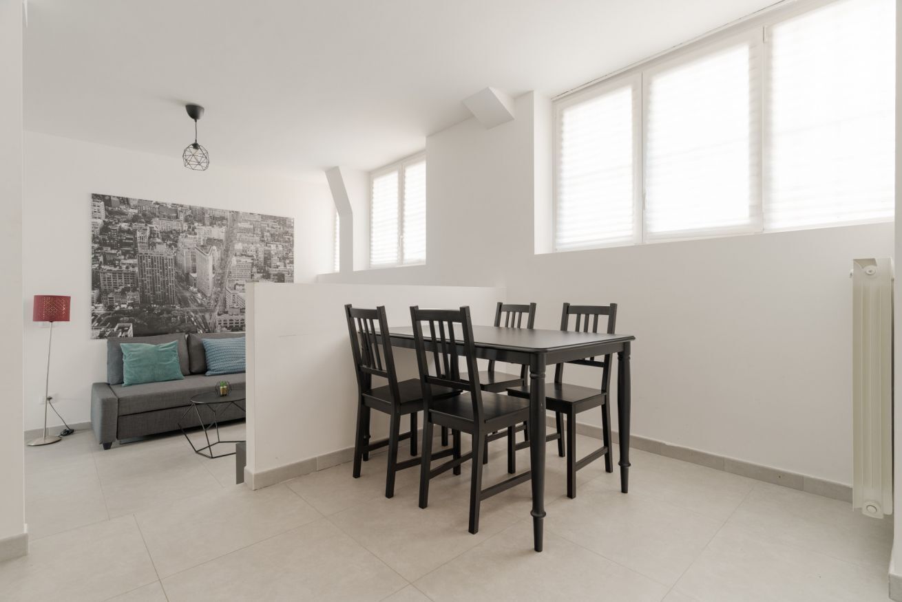A dining area with a table and chairs, a sofa with cushions, a floor lamp, and a cityscape painting on the wall.
