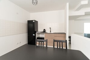 Eine kleine Küche mit einem schwarzen Kühlschrank, einer Mikrowelle und einem Spülbecken. Zwei Holzstühle und eine dunkle Arbeitsplatte sind vorhanden.
