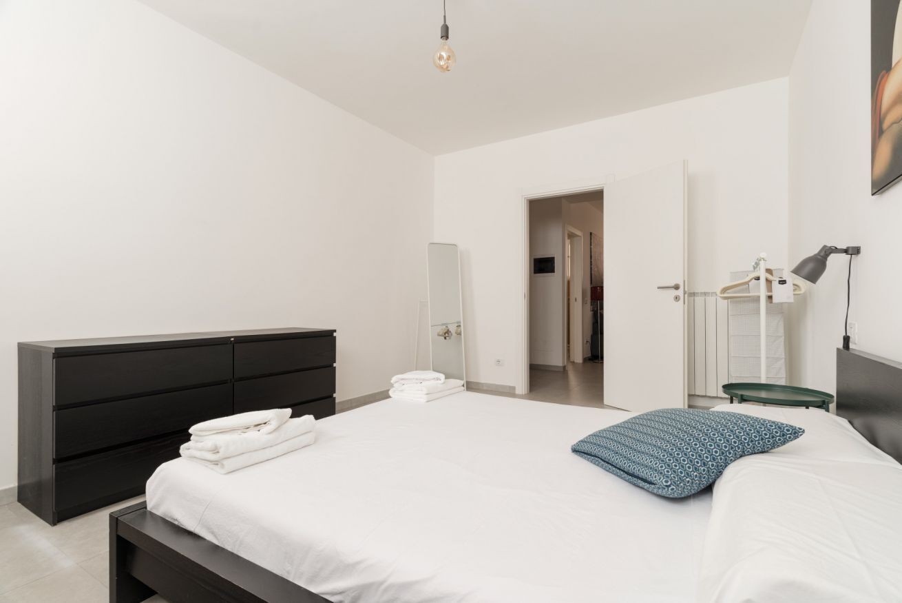 Ein modernes Schlafzimmer mit einem großen Bett, einer dunklen Kommode, einem Spiegel und einer Tür zu einem weiteren Raum.