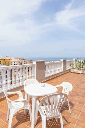 Eine Terrasse mit weißen Plastikstühlen und einem Tisch, mit Blick auf eine Stadtlandschaft und das Meer.