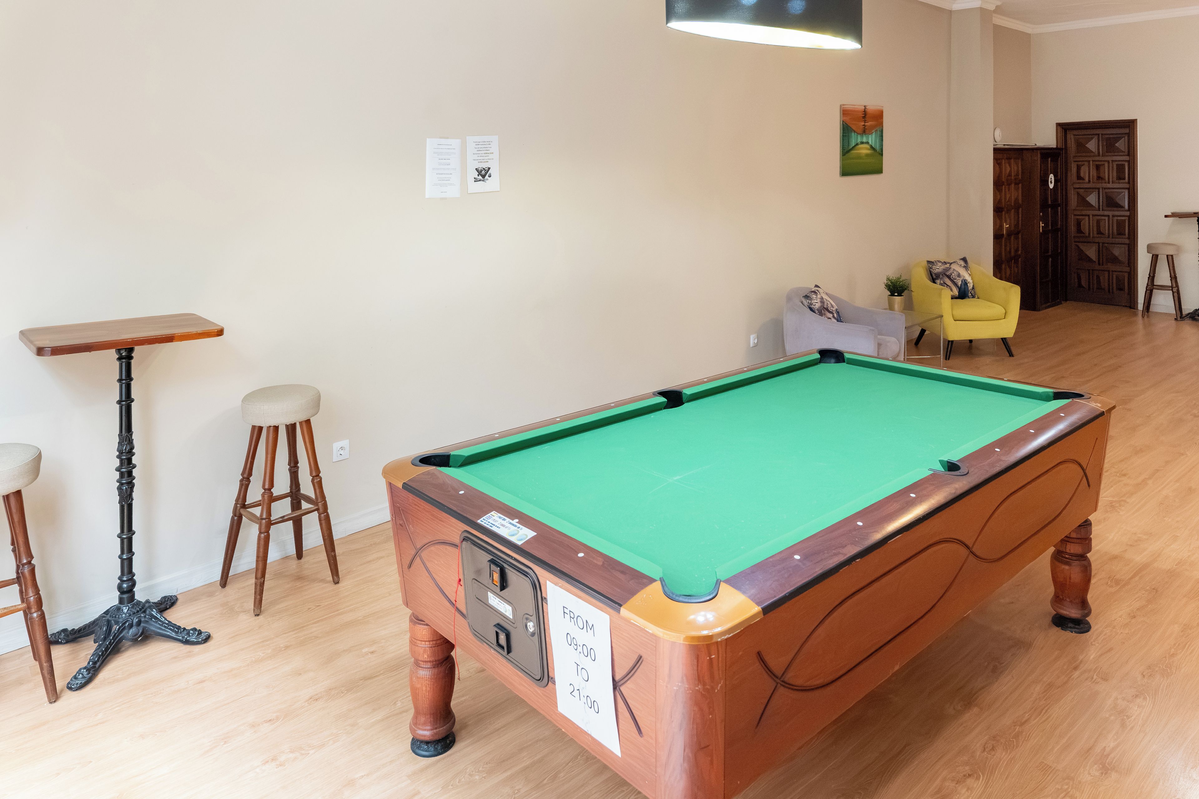 Een biljart in een kamer met houten vloer, een barstoel, een gele fauteuil en een deur op de achtergrond.