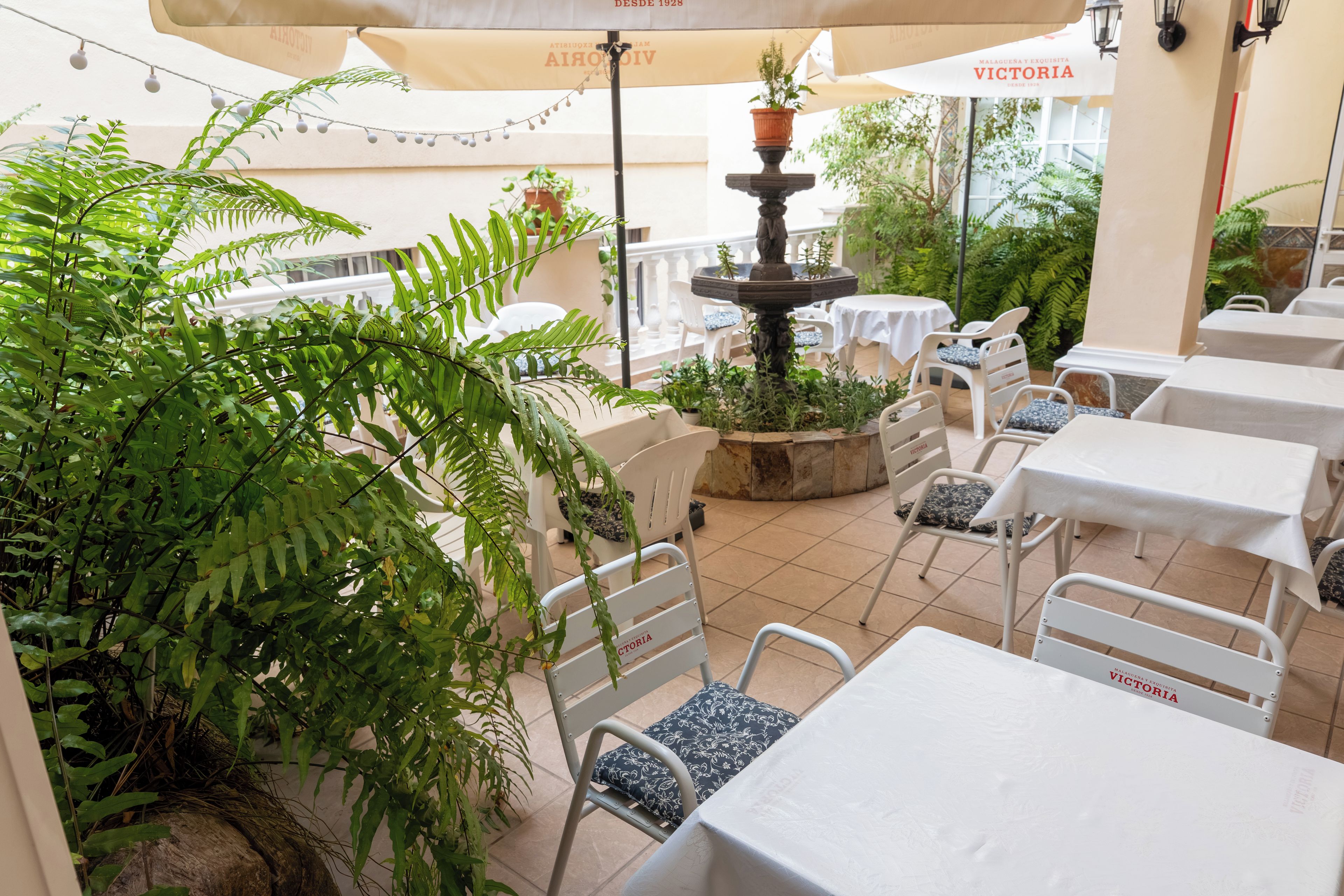 Een terras met tafels en stoelen, omgeven door groen en een centrale fontein.