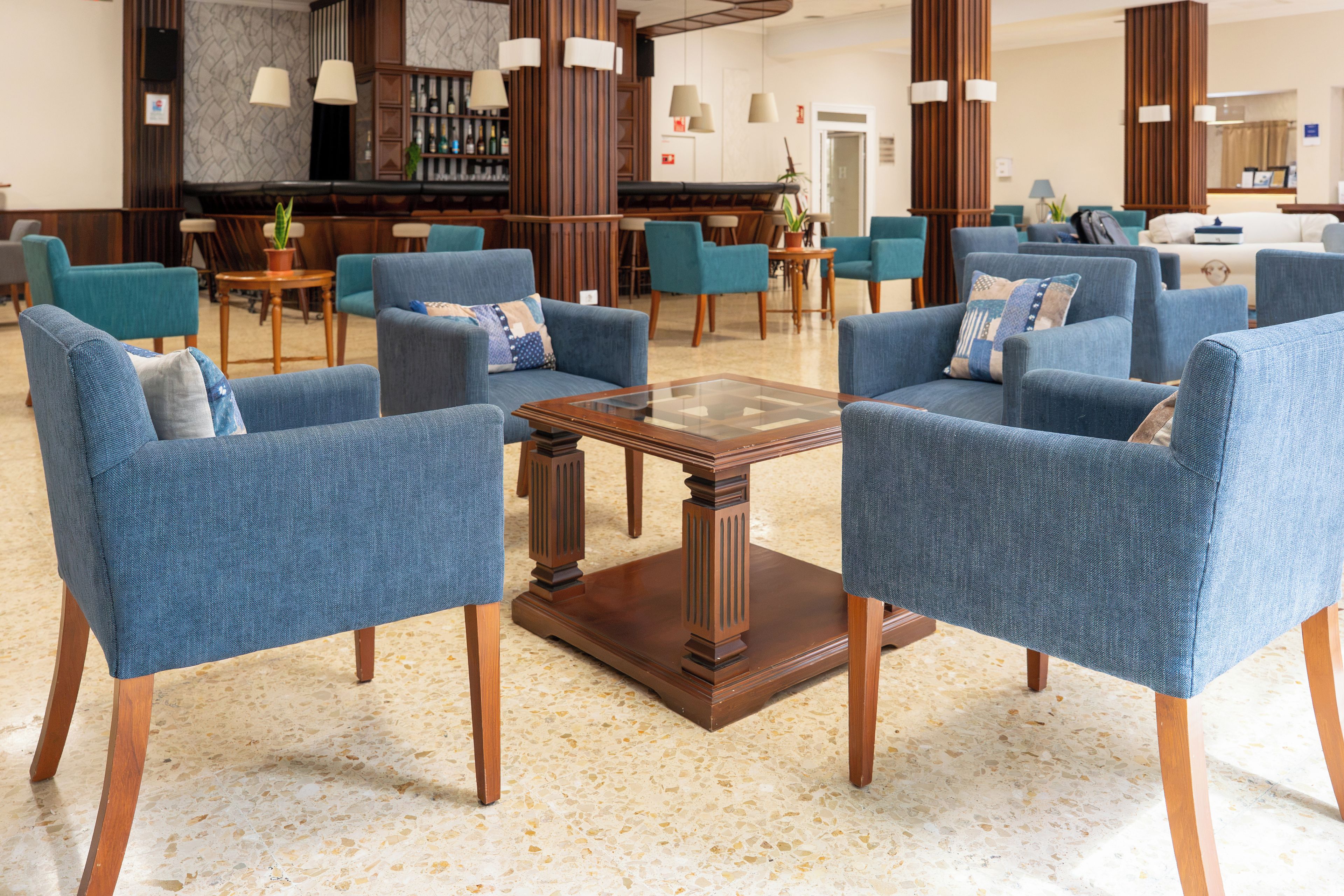 Un vestíbulo con sillones azules dispuestos alrededor de una mesa baja de vidrio central, suelo de madera y una zona de bar al fondo.