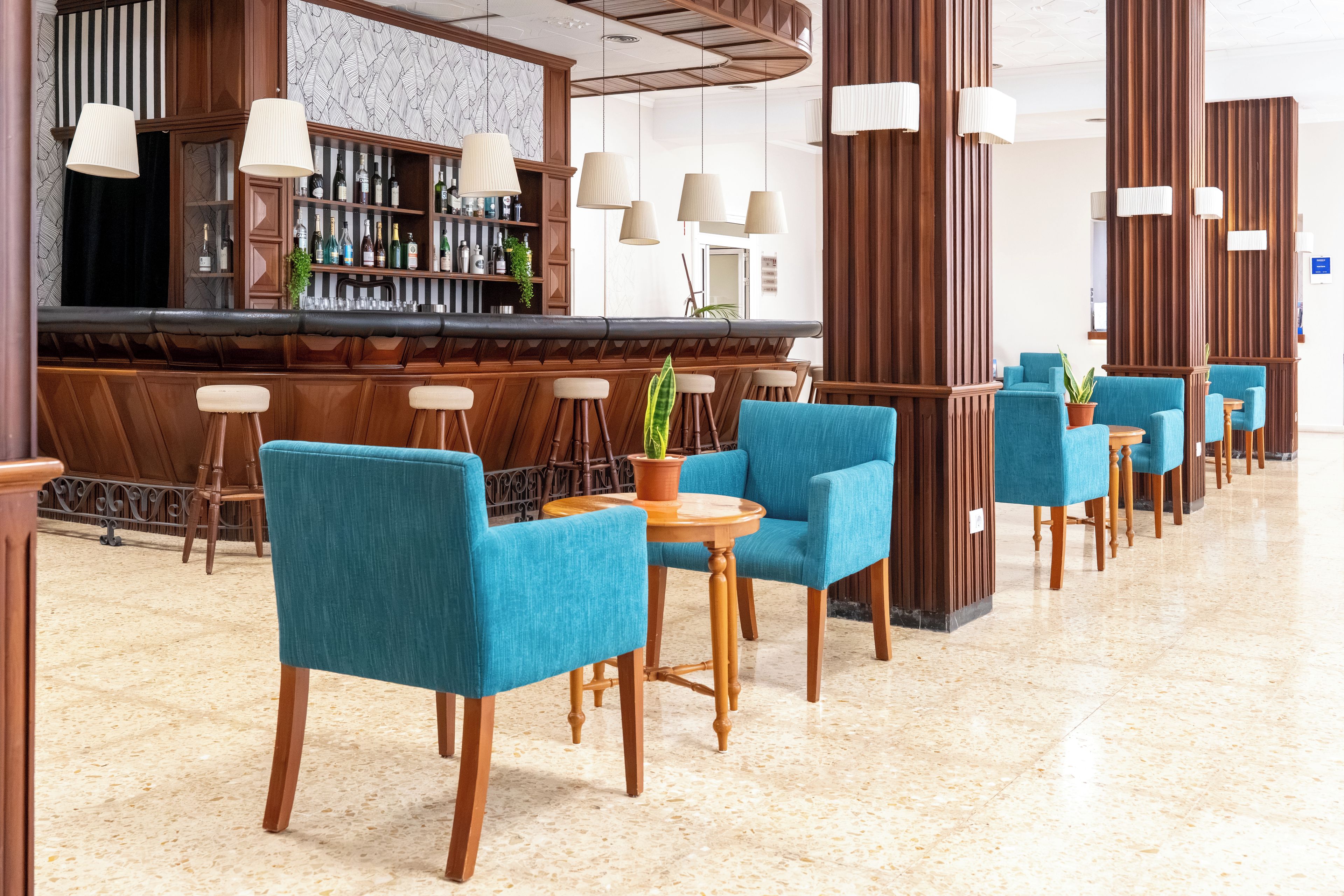 Een bar met een lange toonbank, meerdere hanglampen en een rij blauwe stoelen met houten poten.
