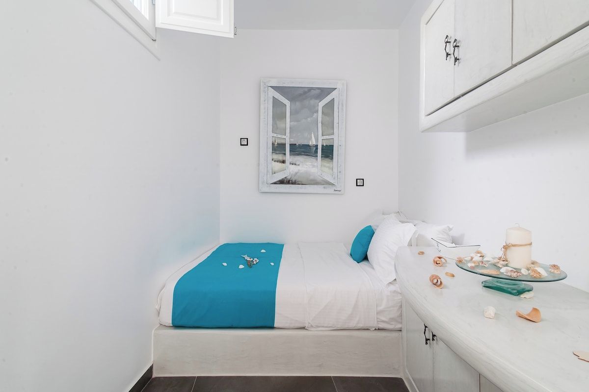 Ein kleiner, weißer Raum mit einem Bett, einem Fenster mit Meerblick und einem Tisch mit Kuchen und Dekoration.