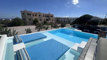 A swimming pool with multiple lanes, surrounded by lounge chairs and umbrellas, with a building in the background.
