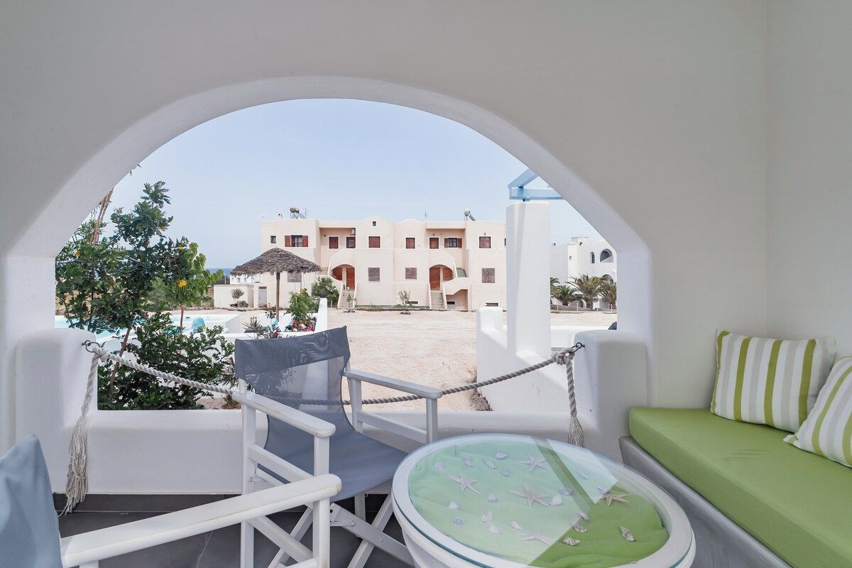 Ein Balkon mit einem weißen Stuhl, einem runden Tisch mit grüner Tischdecke und Blick auf ein Gebäude mit Bögen und einem Pool.