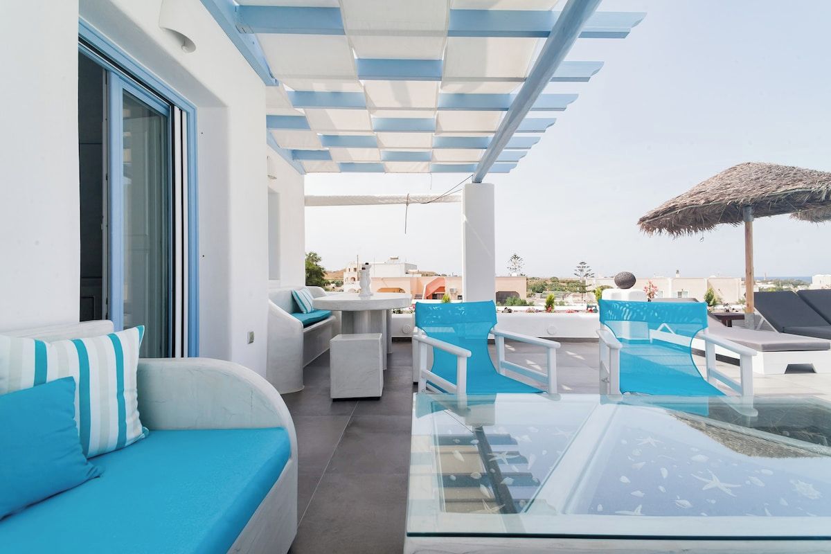 Eine Dachterrasse mit einem Glastisch, blauer und weißer Möbel und einem Reetdachschirm.