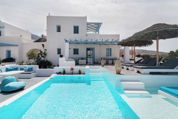 A pool area with a modern white building, outdoor seating, and a thatched umbrella.