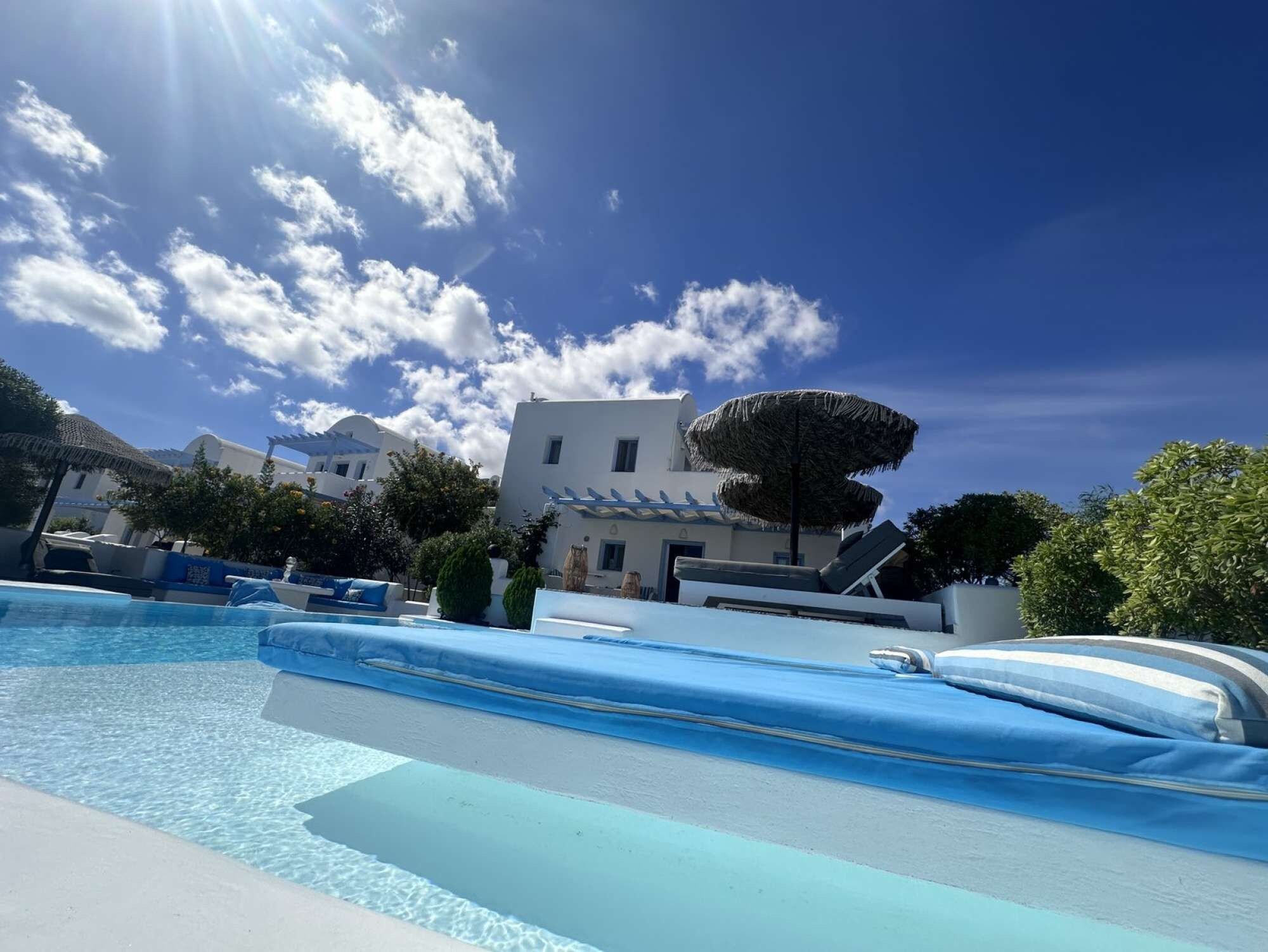 Ein Poolbereich mit klarem, blauem Wasser, ein weißes Gebäude mit markanter Architektur und eine große Sonnenschirmkonstruktion.