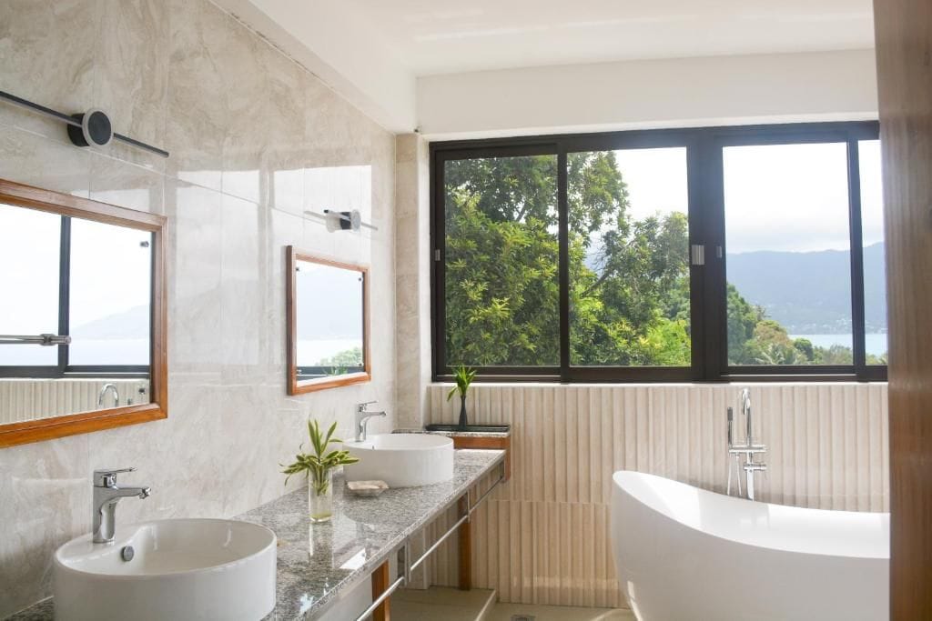 A modern bathroom with a freestanding bathtub, a double sink vanity, and a large window offering a view of greenery and mountains.