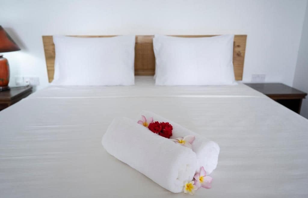Una cama bien hecha con una toalla enrollada y flores, un cabecero de madera y una mesita de noche con una lámpara.