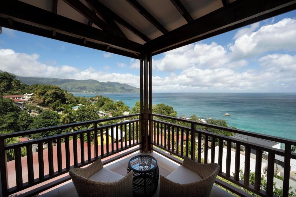 A balcony with wicker furniture and a glass table, overlooking a coastal landscape with mountains and a clear sky.
